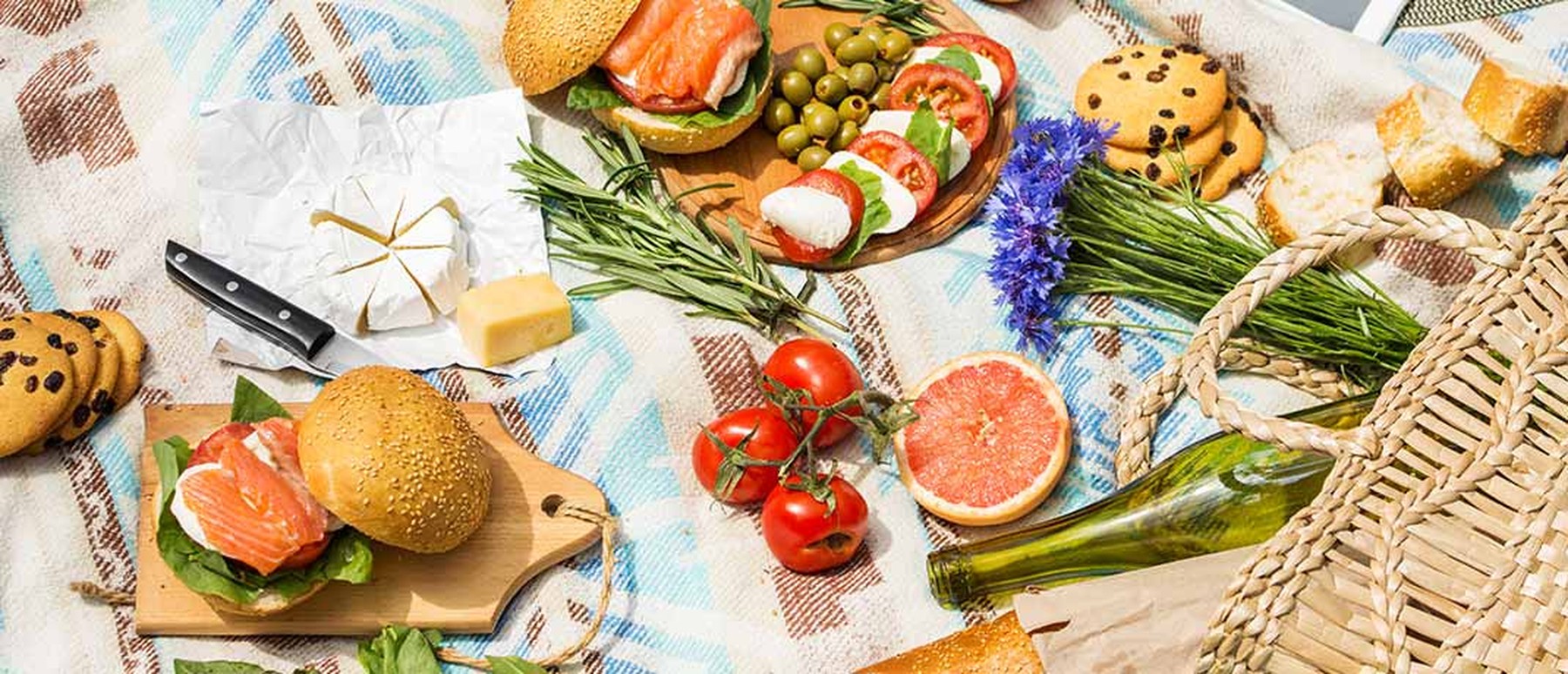 picnic setting on grass covered with a blanket with picnic food across it such as grapefruit, cookies, salmon bagel and tomatoes 