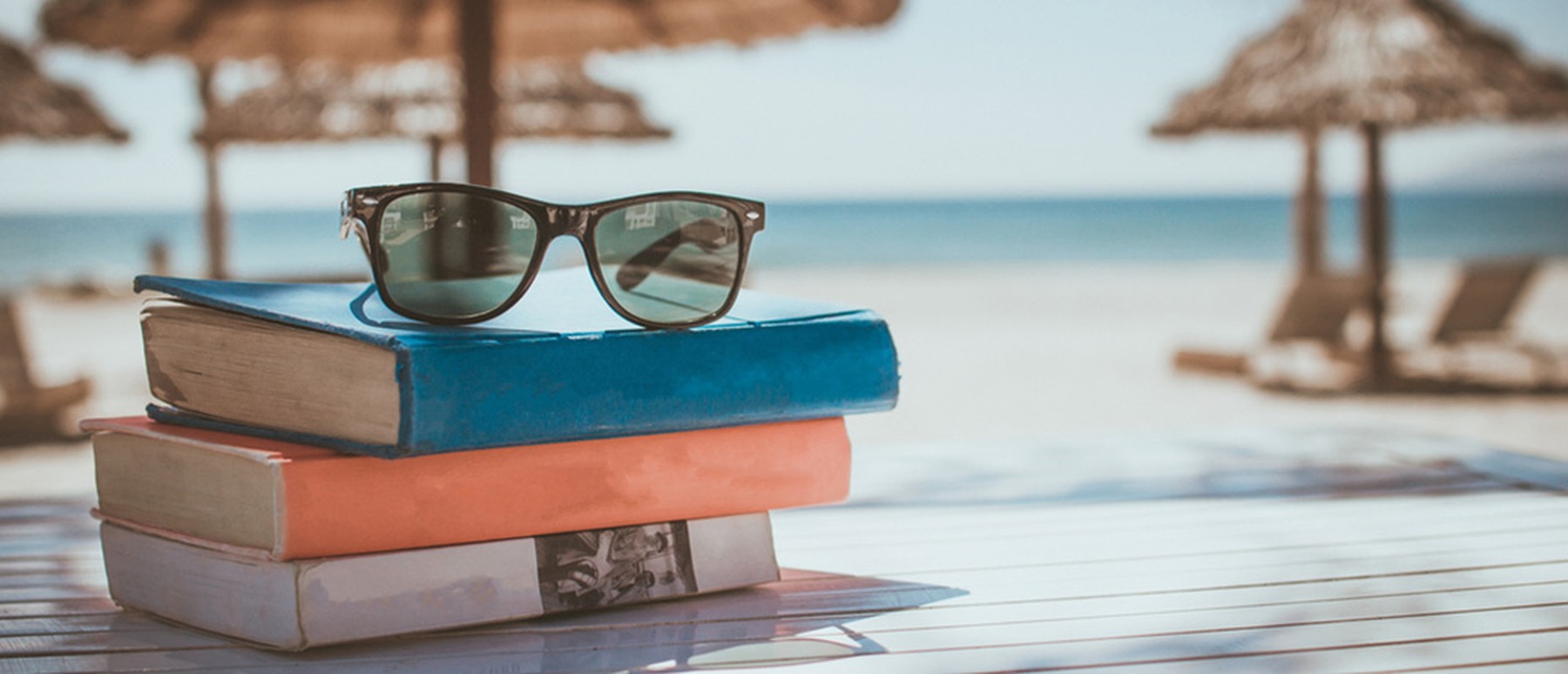 Bücher und Sonnenbrille gelegt auf einem Tisch und Sonnenschirme und Strand im Hintergrund